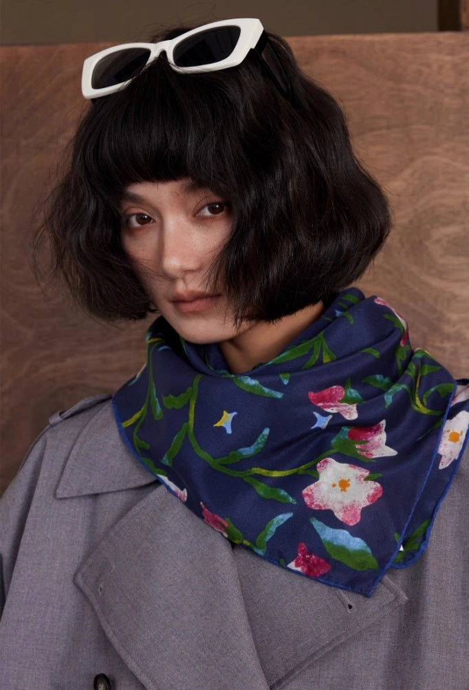 Woman wearing a Midnight Blue silk square scarf (63x63cm) as a neck accessory over a gray coat, featuring a floral mulberry silk pattern, with white sunglasses and a wooden background.