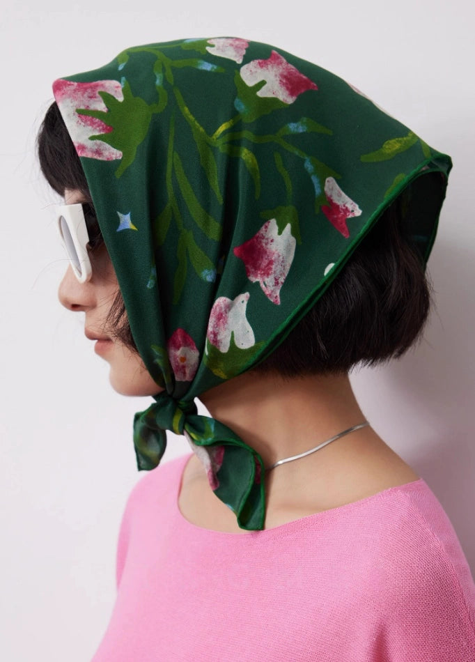 Side profile of a woman wearing a Wilderness Green silk square scarf (63x63cm) as a headscarf, featuring pink morning glory floral pattern on mulberry silk, with white sunglasses and a pink top.