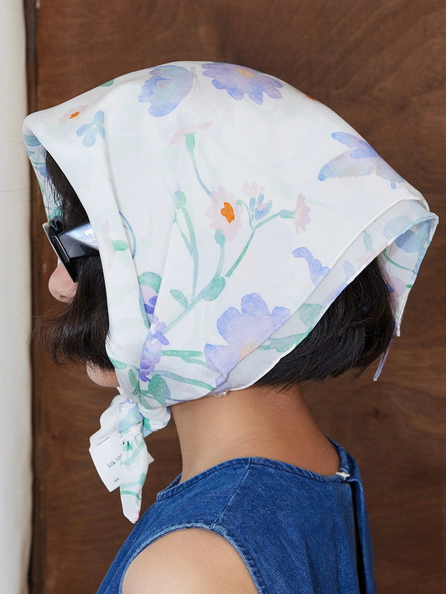 A square silk scarf with a watercolor floral design worn as a headscarf, close-up emphasizing how it is styled.